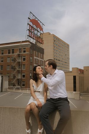 A couple in white and dark attire shares tender moments on a city rooftop under cloudy skies.