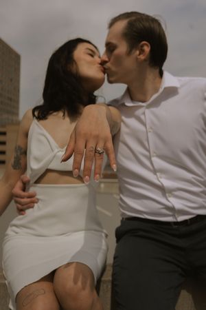A romantic couple in white and dark clothing shows affection while sitting on an urban ledge.