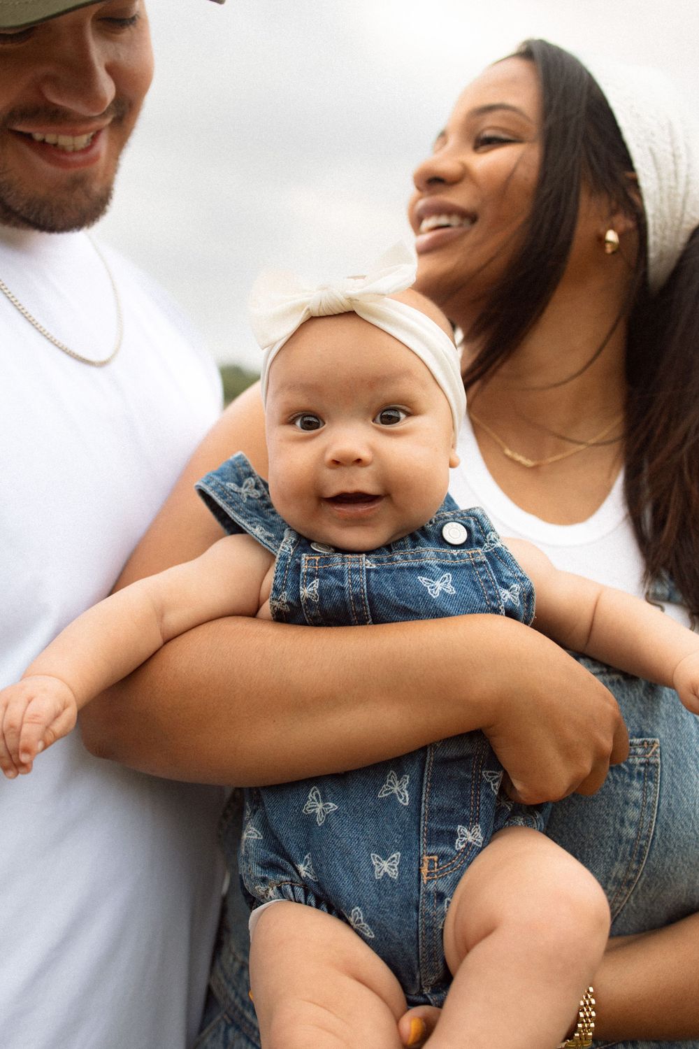 Baby wearing denim overalls and white headband smiles while being held in a loving family embrace outdoors.