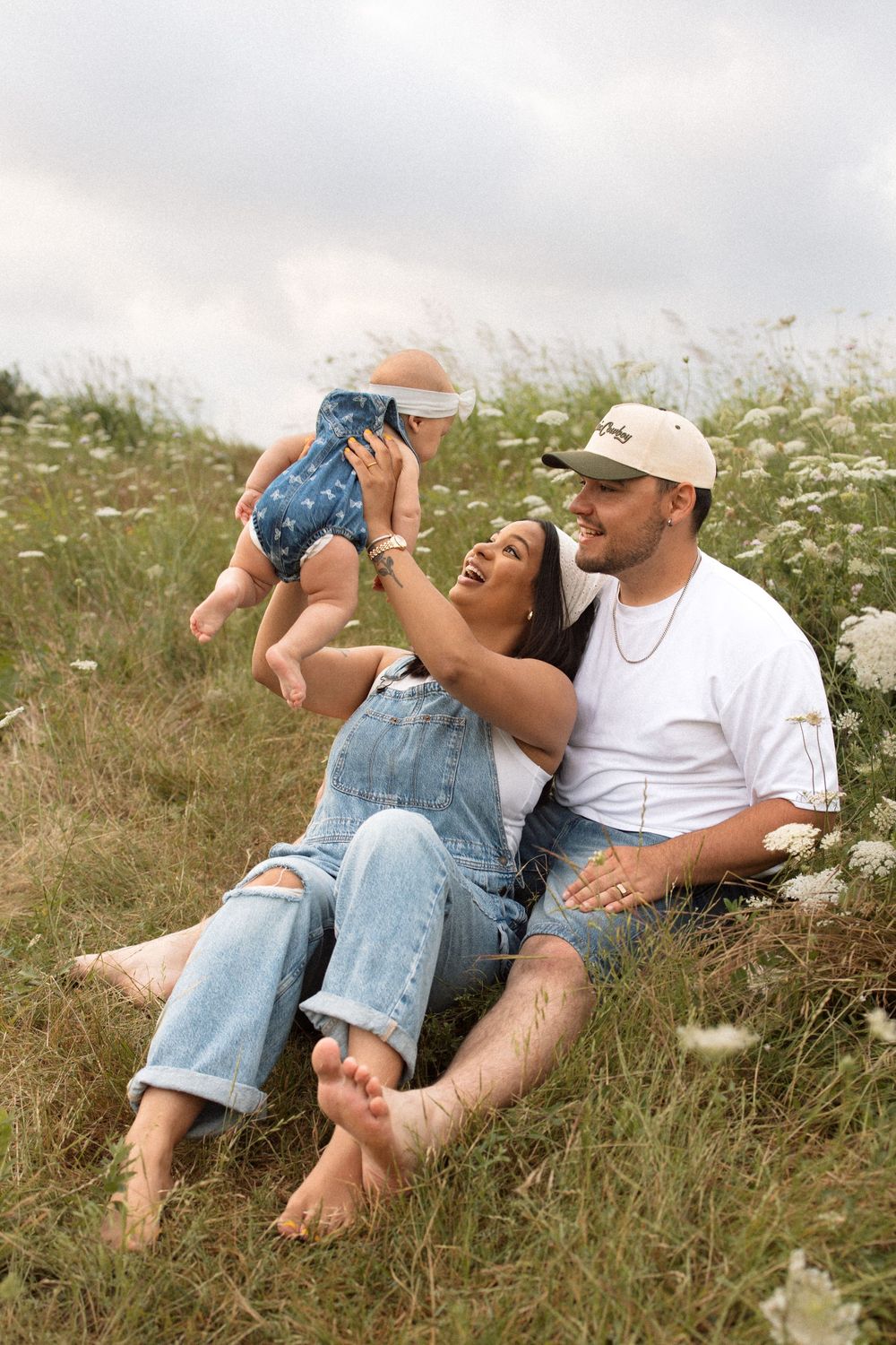 Joyful young couple sitting in wildflower meadow with a baby wearing blue romper on a summer day.
