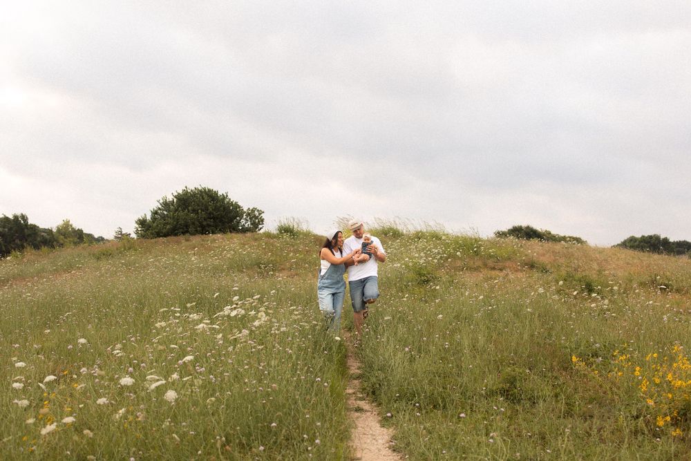 A couple embraces while standing on a wildflower-dotted dirt path through rolling green hills on a cloudy day.