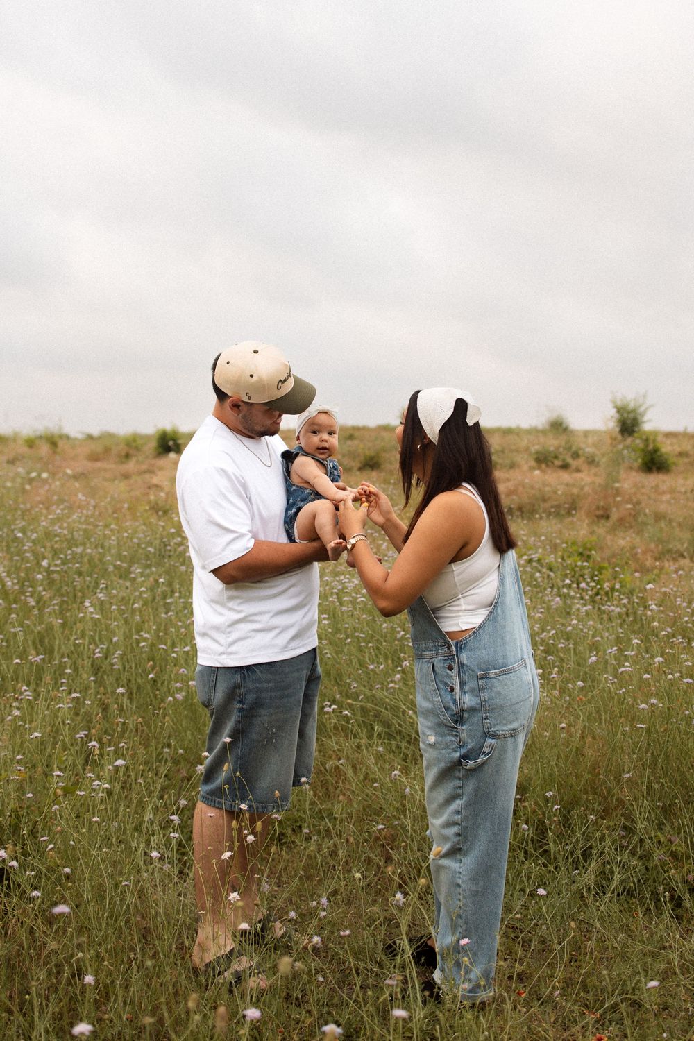 An adult couple stands together in a wildflower field, wearing casual attire and holding a small baby on a cloudy day.