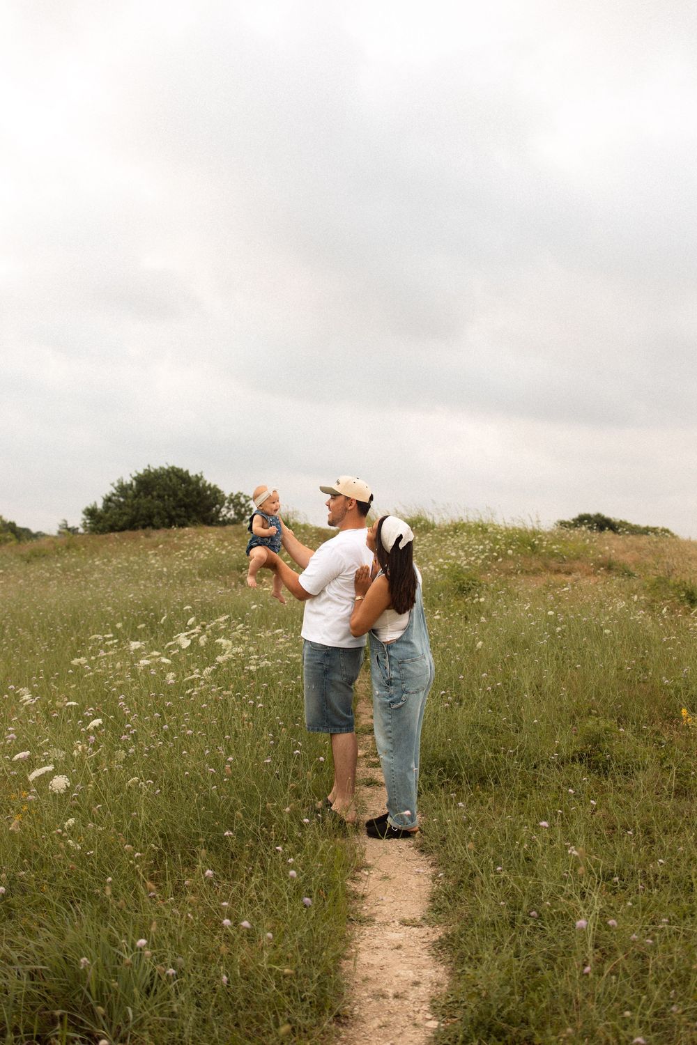 A couple walking on a dirt path through a wildflower meadow lifting a small child against an overcast sky.