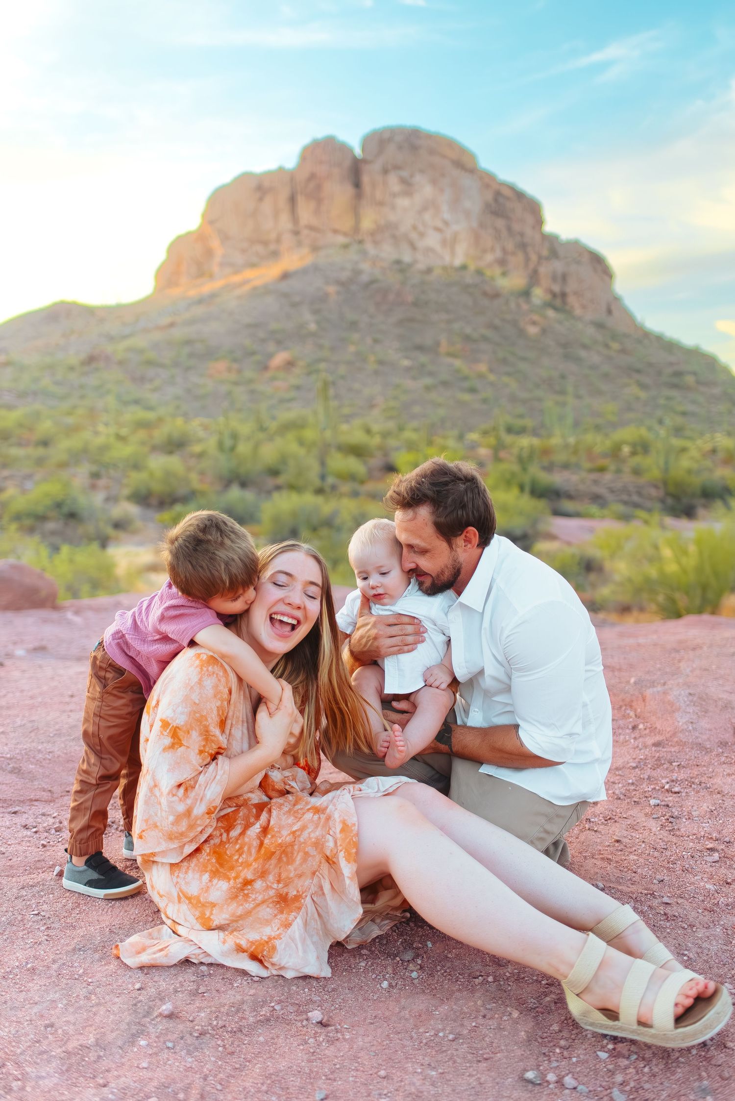 Family sits together laughing on red desert ground with dramatic mountain formation in background at sunset.