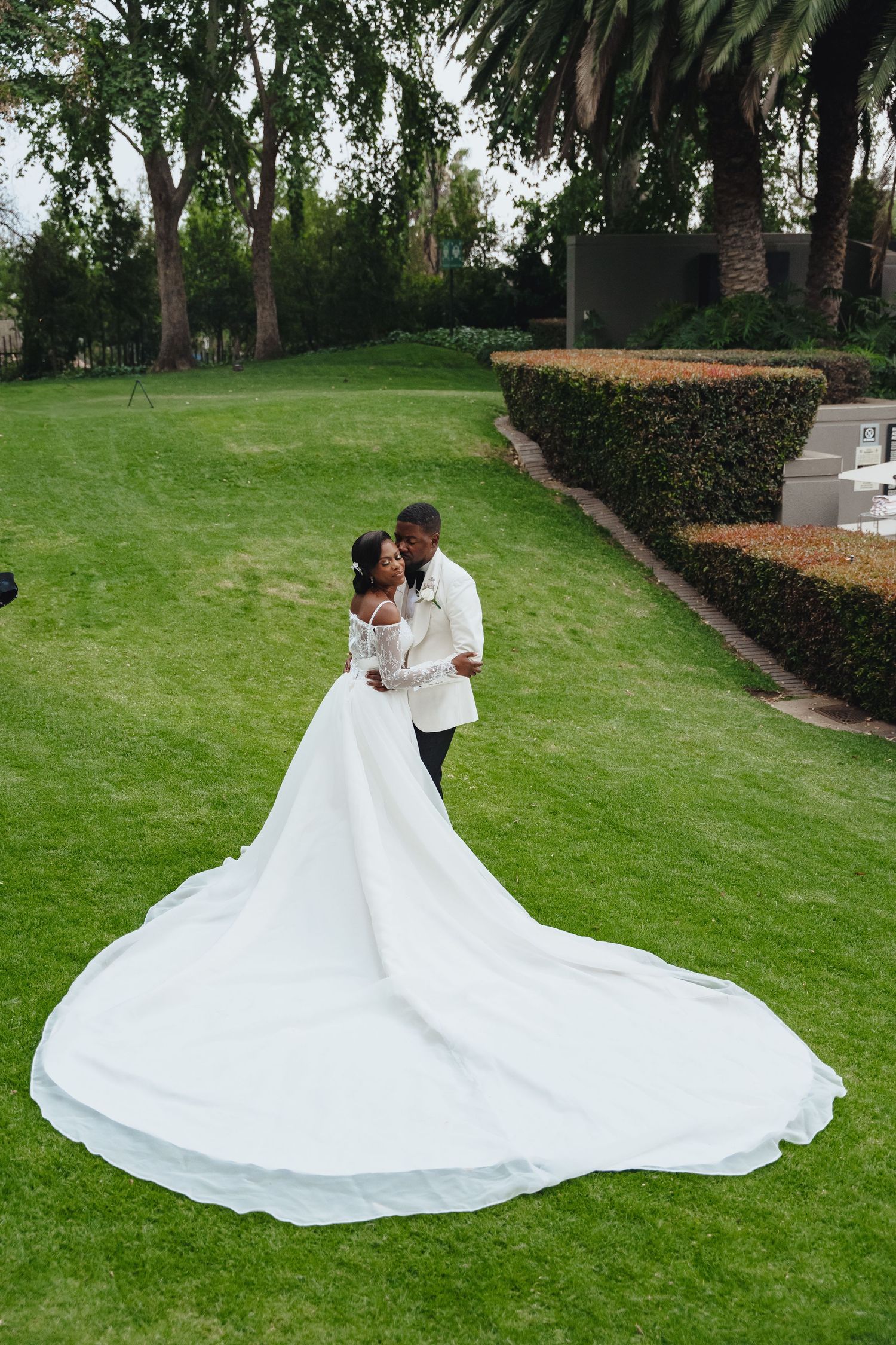 A romantic wedding couple shares tender moments on a lush green lawn surrounded by palm trees and manicured landscaping.