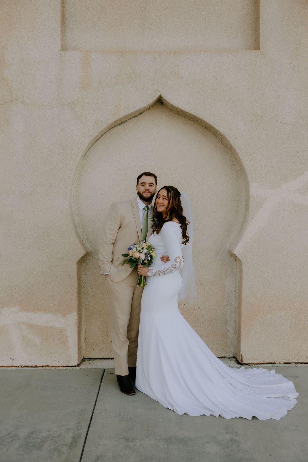 A couple poses in traditional wedding attire against an ornate archway in soft lighting.