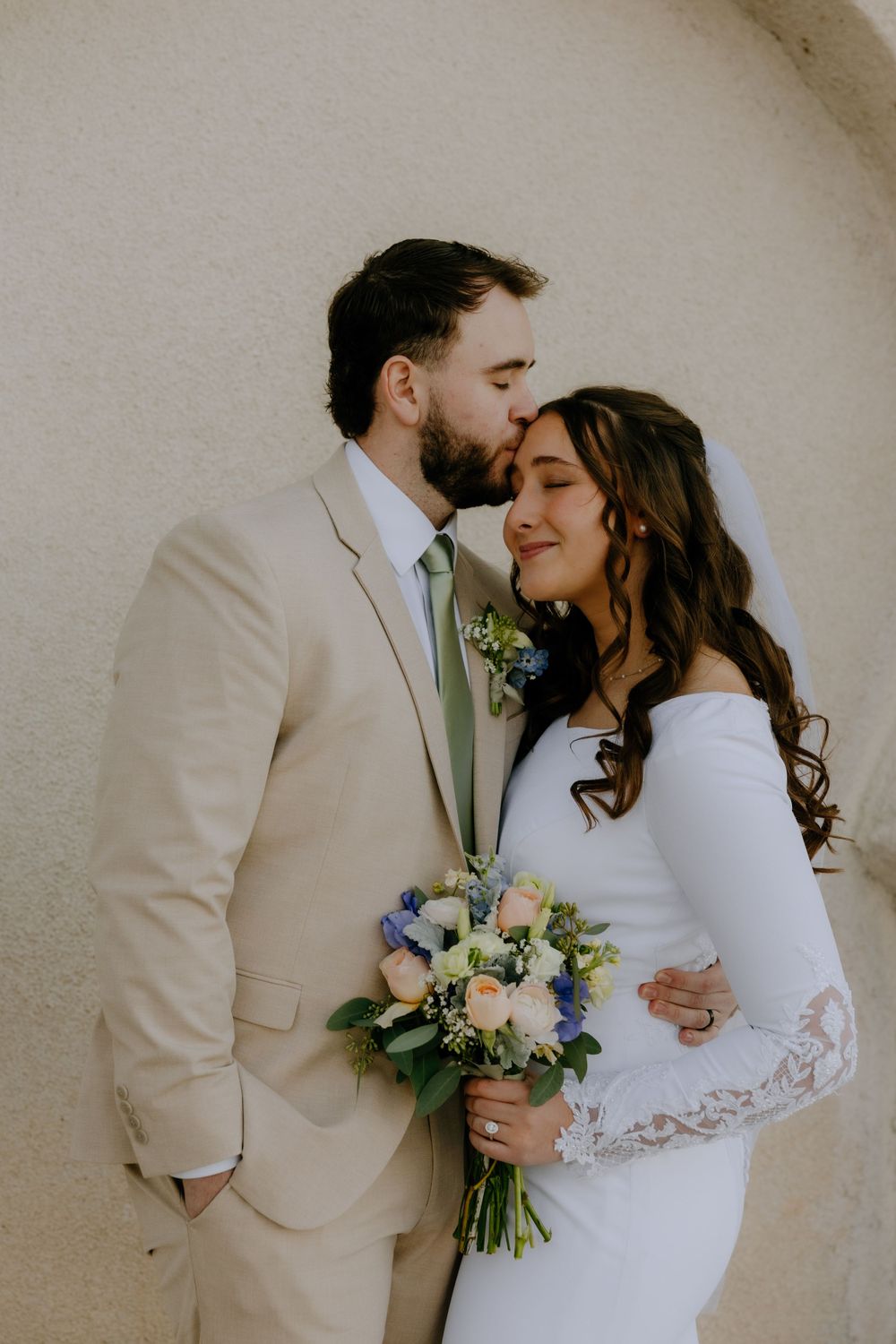 A romantic wedding couple in tan suit and white dress share an intimate moment against a light wall with floral bouquet.