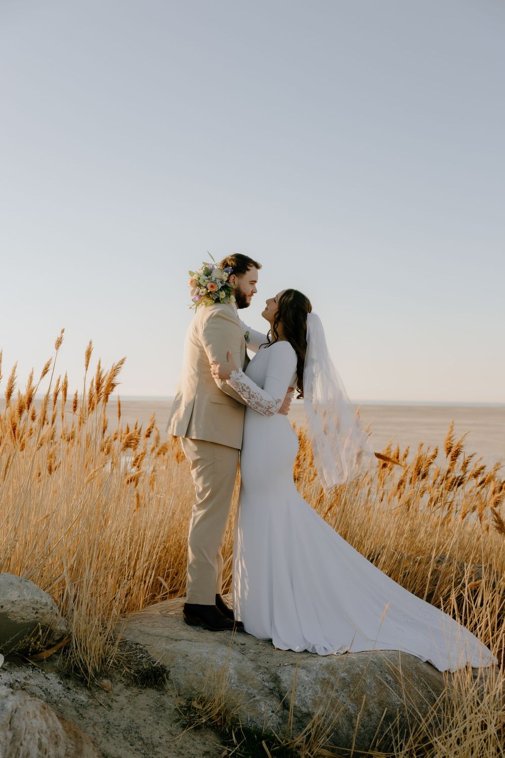A romantic wedding couple stands among tall wheat grass at sunset against a soft sky backdrop.