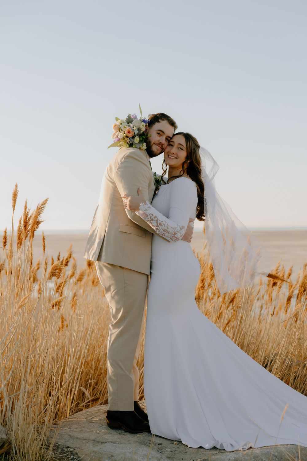 A wedding couple embraces in a field of golden wheat during sunset.