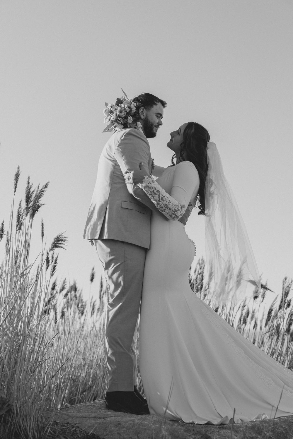 Black and white photo of wedding couple standing in tall grass with flowing veil.