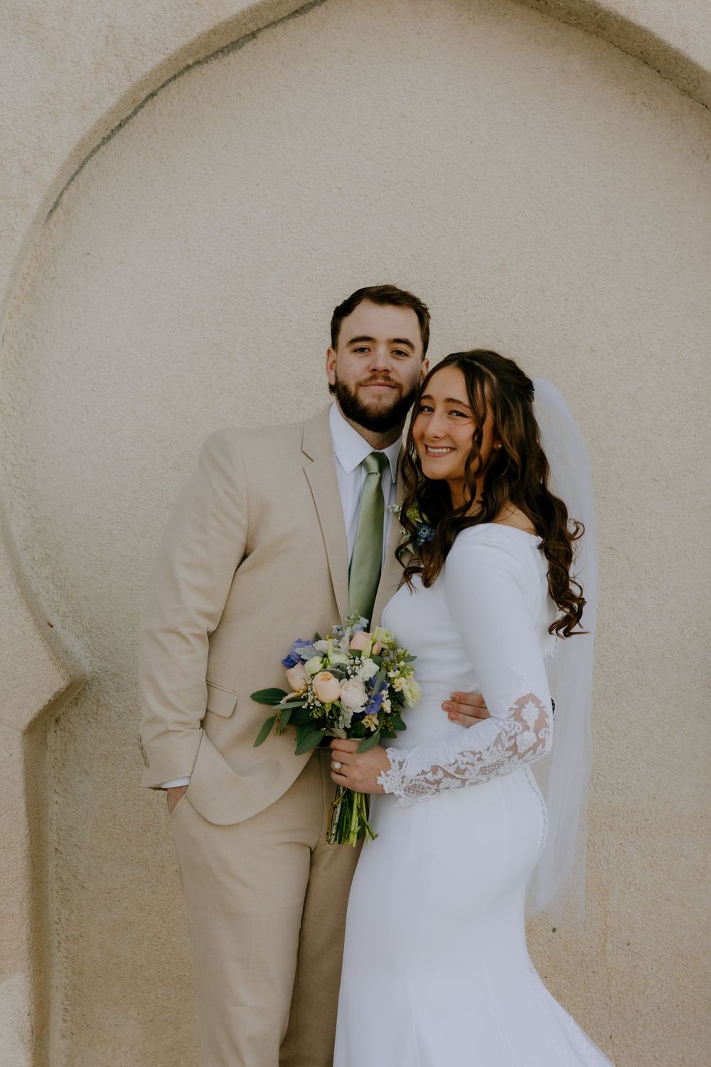A bride and groom pose together against a pale wall, with the bride holding a beautiful floral bouquet.