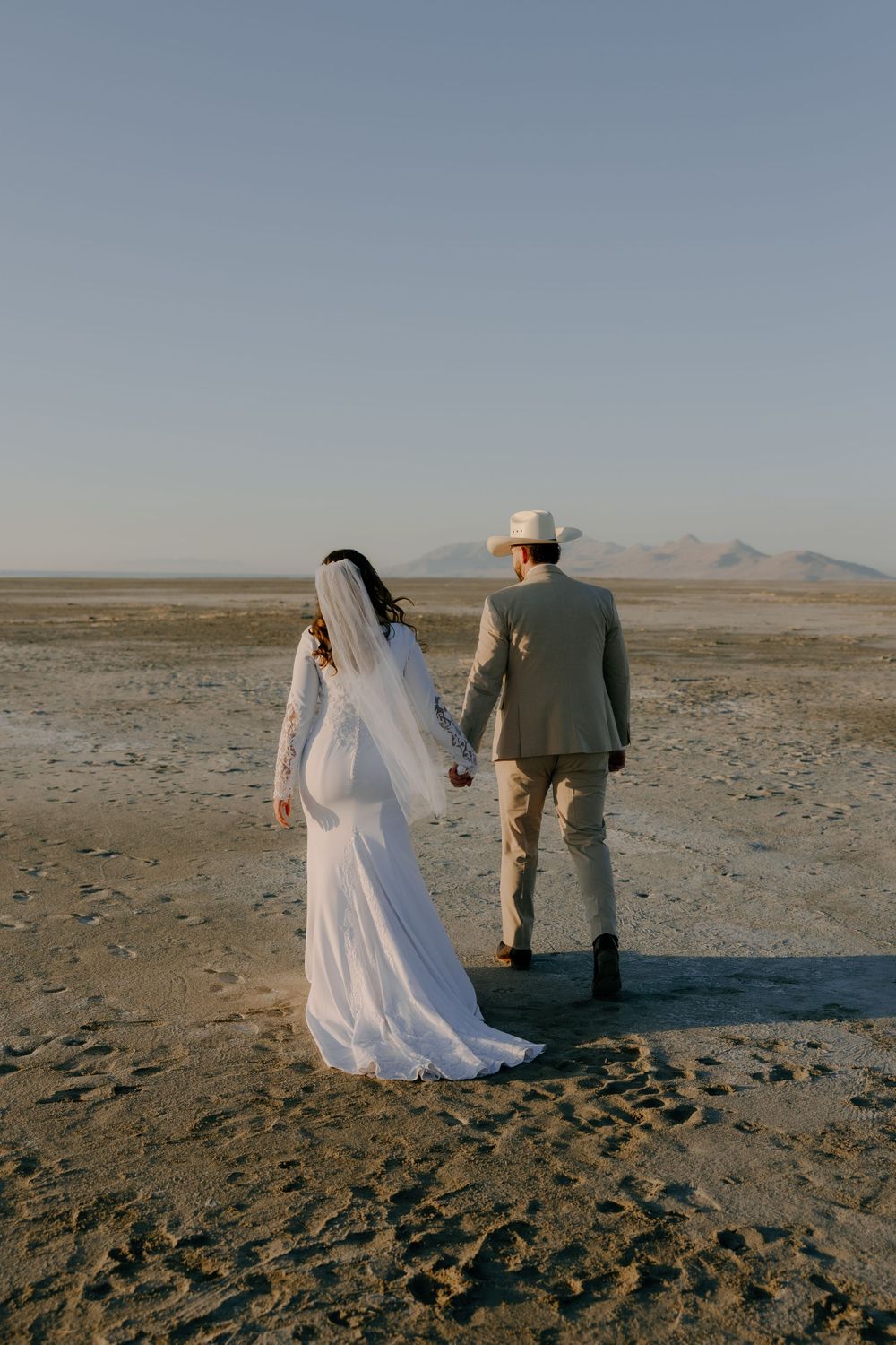 Newlywed couple walks hand in hand across a desert landscape with mountains in the distance at sunset.