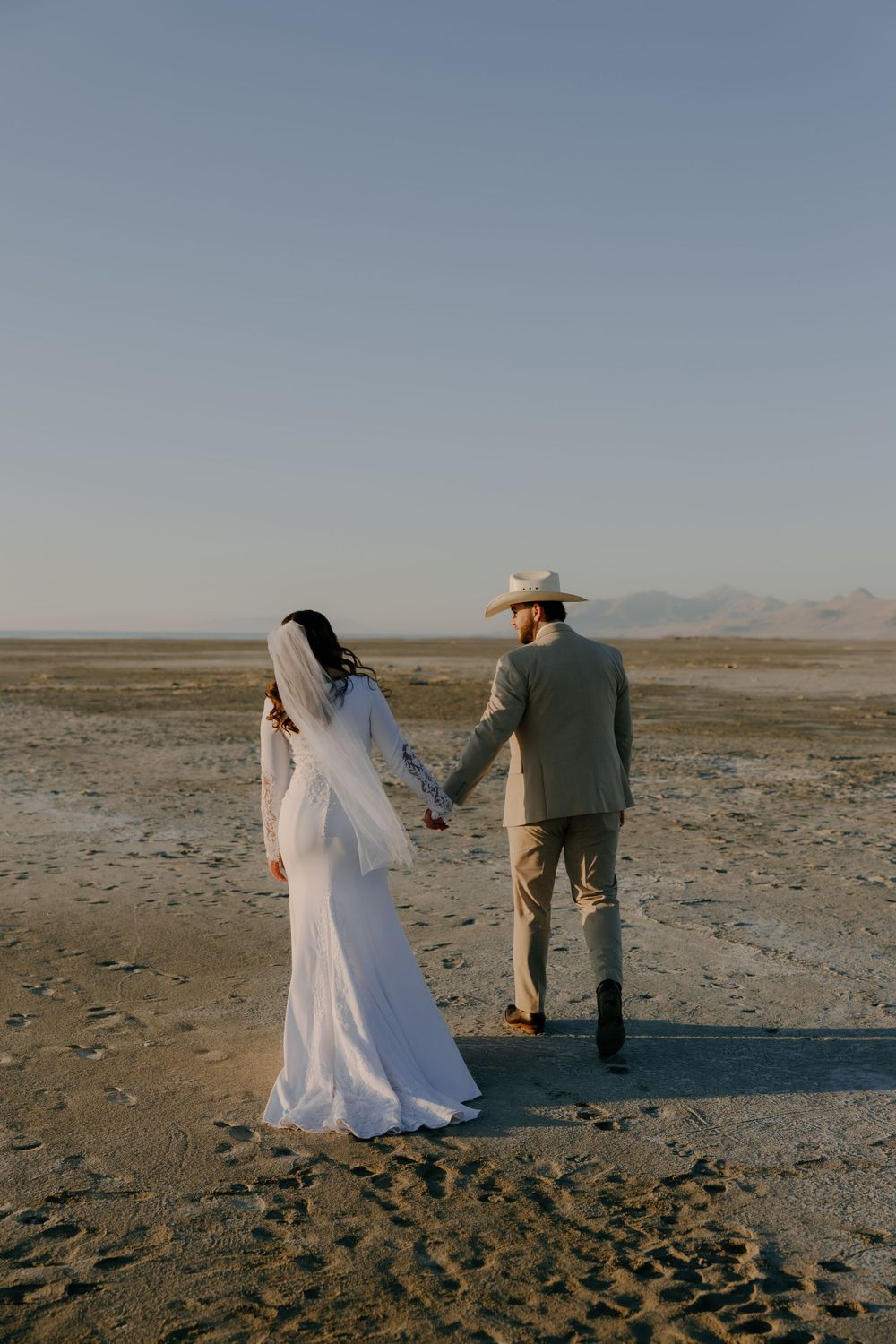 A couple in wedding attire hold hands while standing on a vast desert plain at sunset with mountains in the distance.