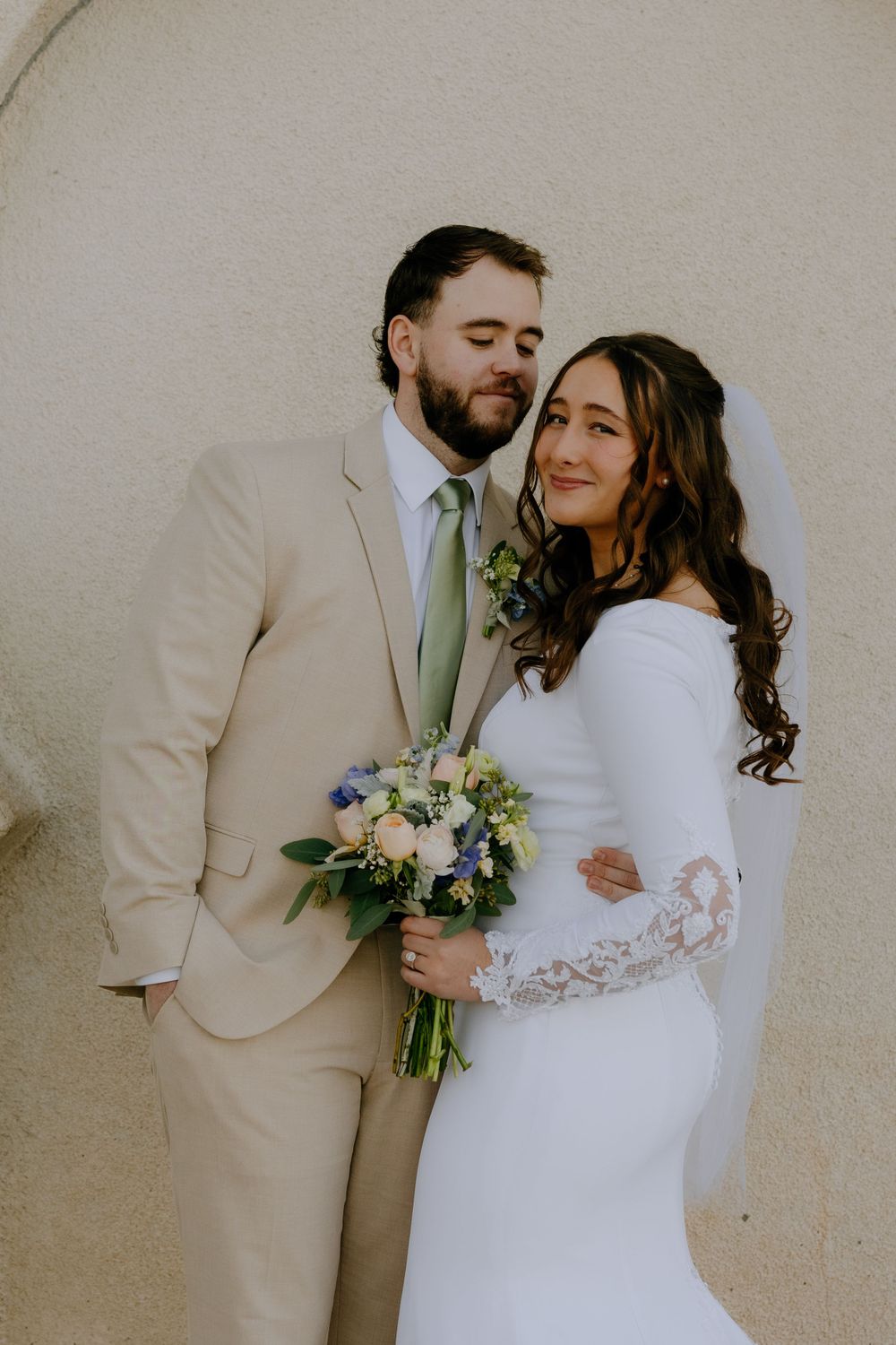 A romantic wedding couple poses together against a neutral wall, with bride in white lace gown holding a blue bouquet.
