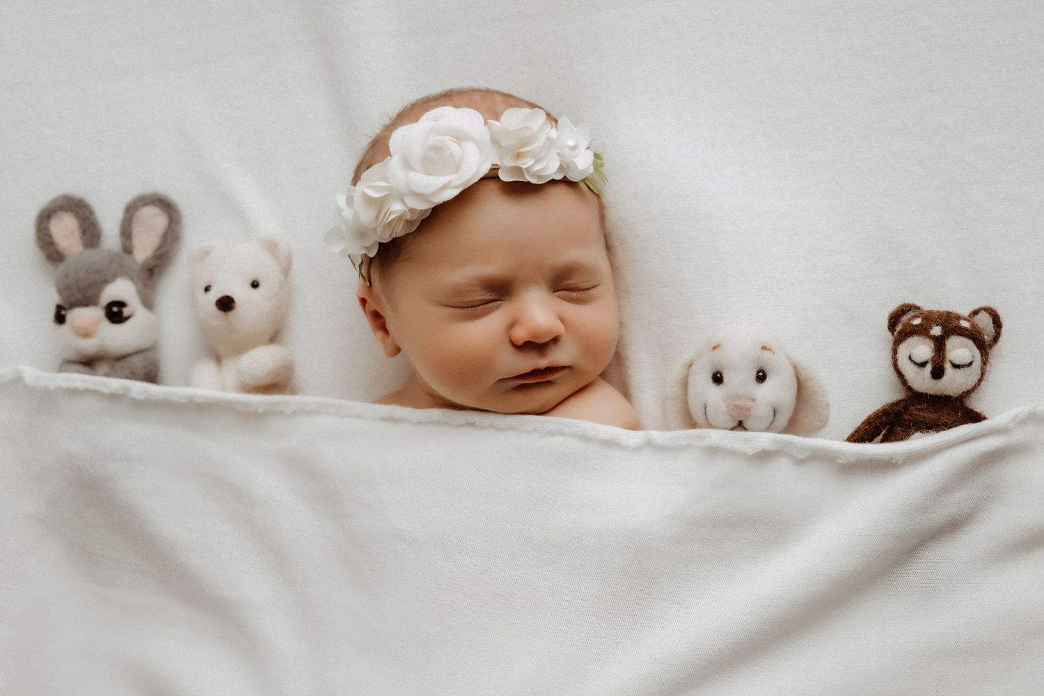 A sleeping infant rests on white bedding surrounded by stuffed animals including a bunny and teddy bear.