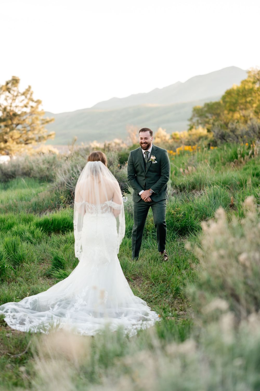 A couple shares an emotional first look moment on a hillside with lush green grass and mountains in the background.