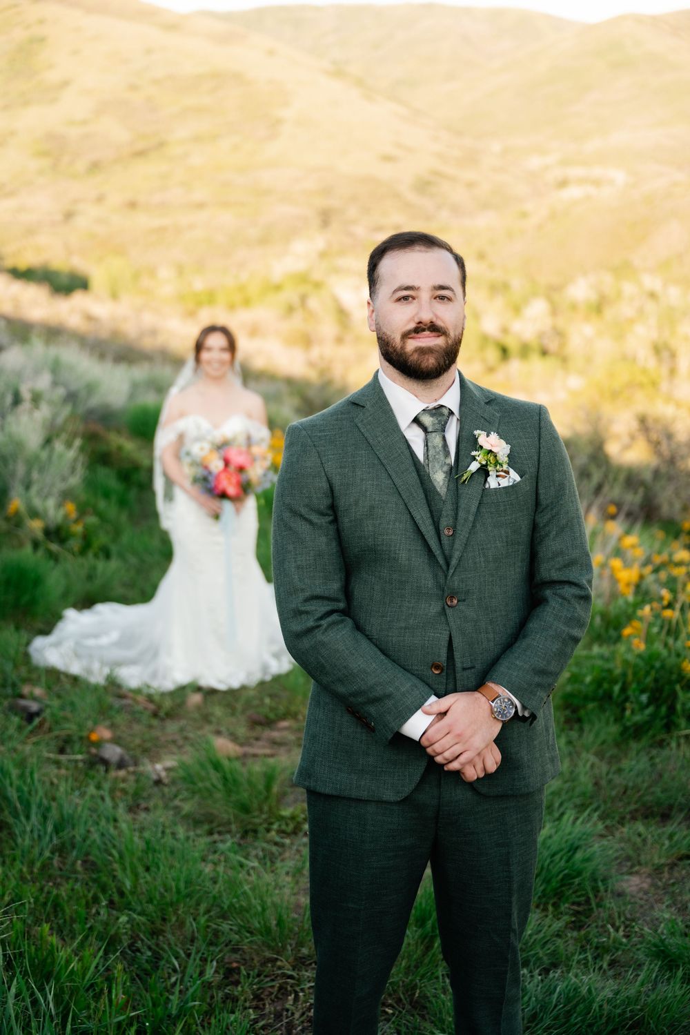 A couple in wedding attire poses on a hillside during golden hour with mountains in the background.