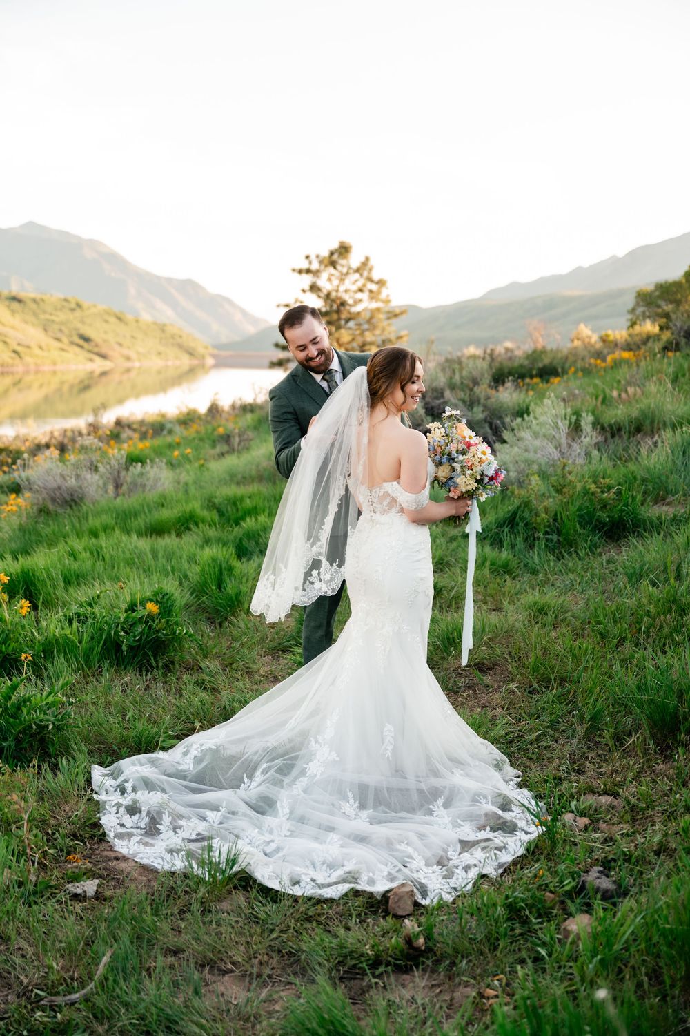 Couple shares intimate moment on grassy hillside overlooking lake with flowery bouquet and long lace wedding train.