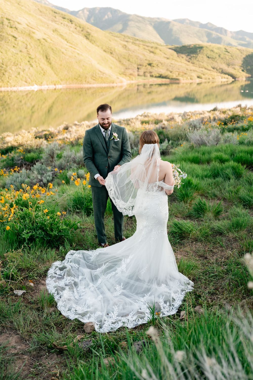A flowing white wedding dress train trails across mountain wildflowers as couple poses near a scenic lake vista.