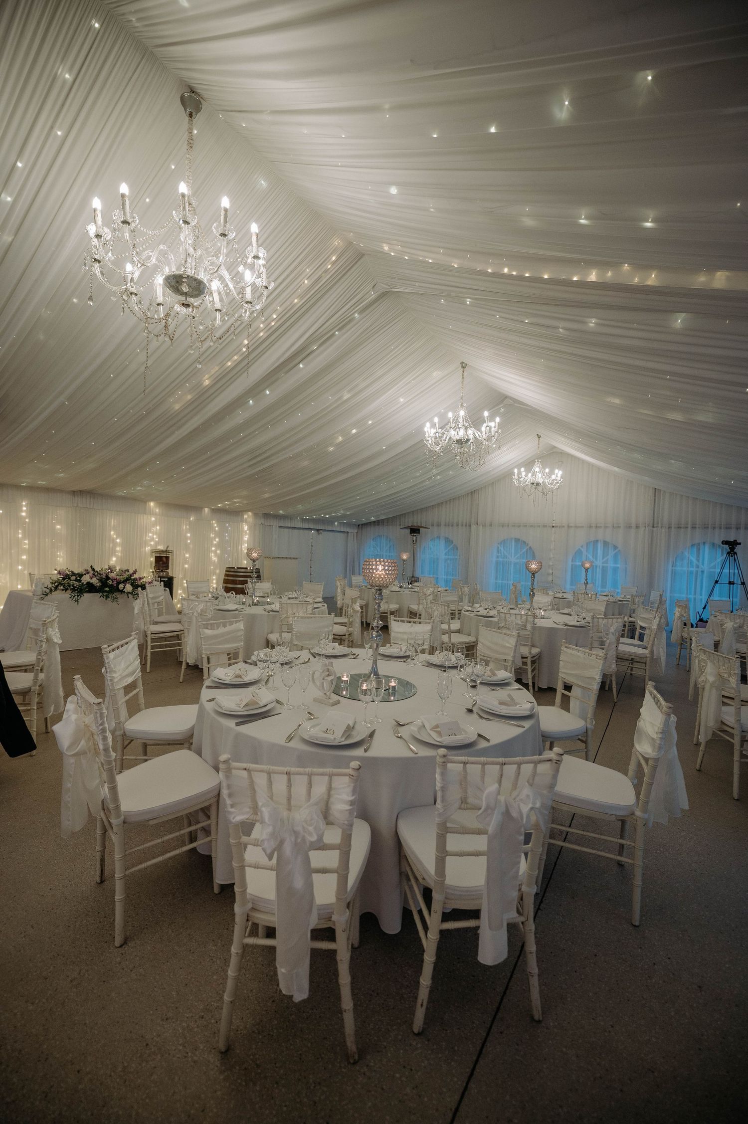Elegant white wedding reception tent with draped fabric ceiling, crystal chandeliers, and round tables set for dinner service.