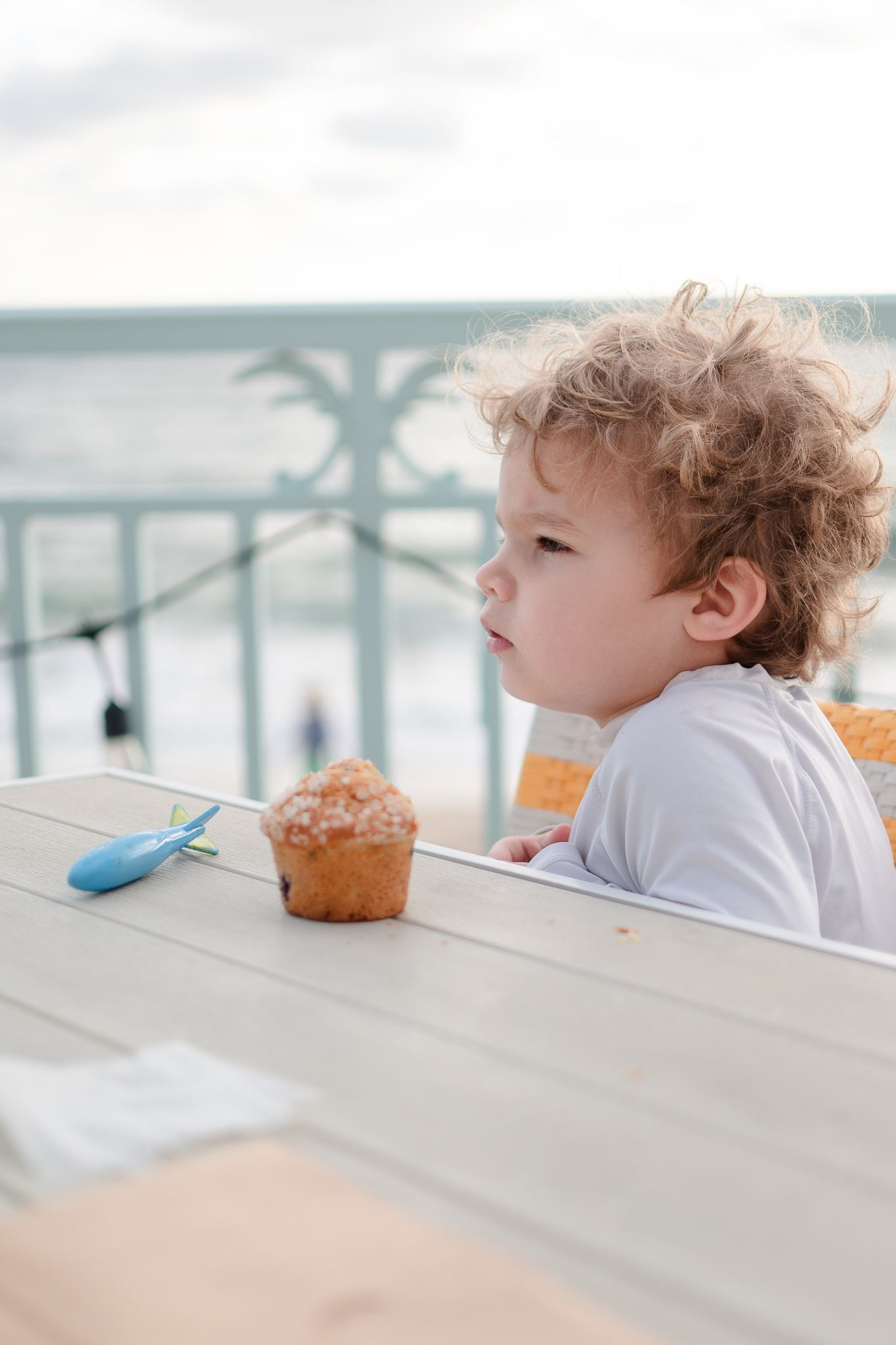 A small child with curly hair sits at an outdoor table with a muffin, overlooking a waterfront view.