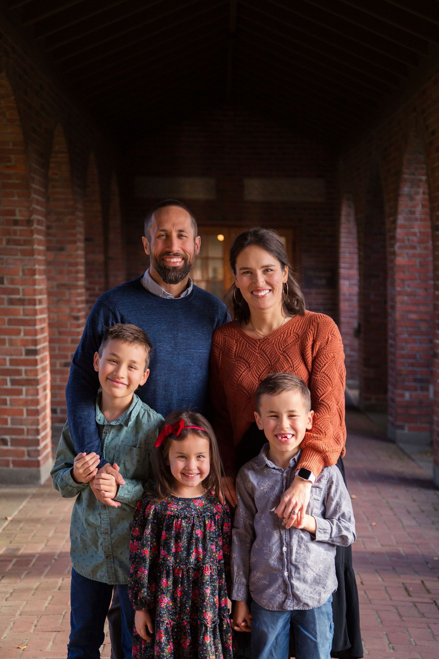 A family poses together under a brick archway corridor with natural lighting falling on their smiling faces.