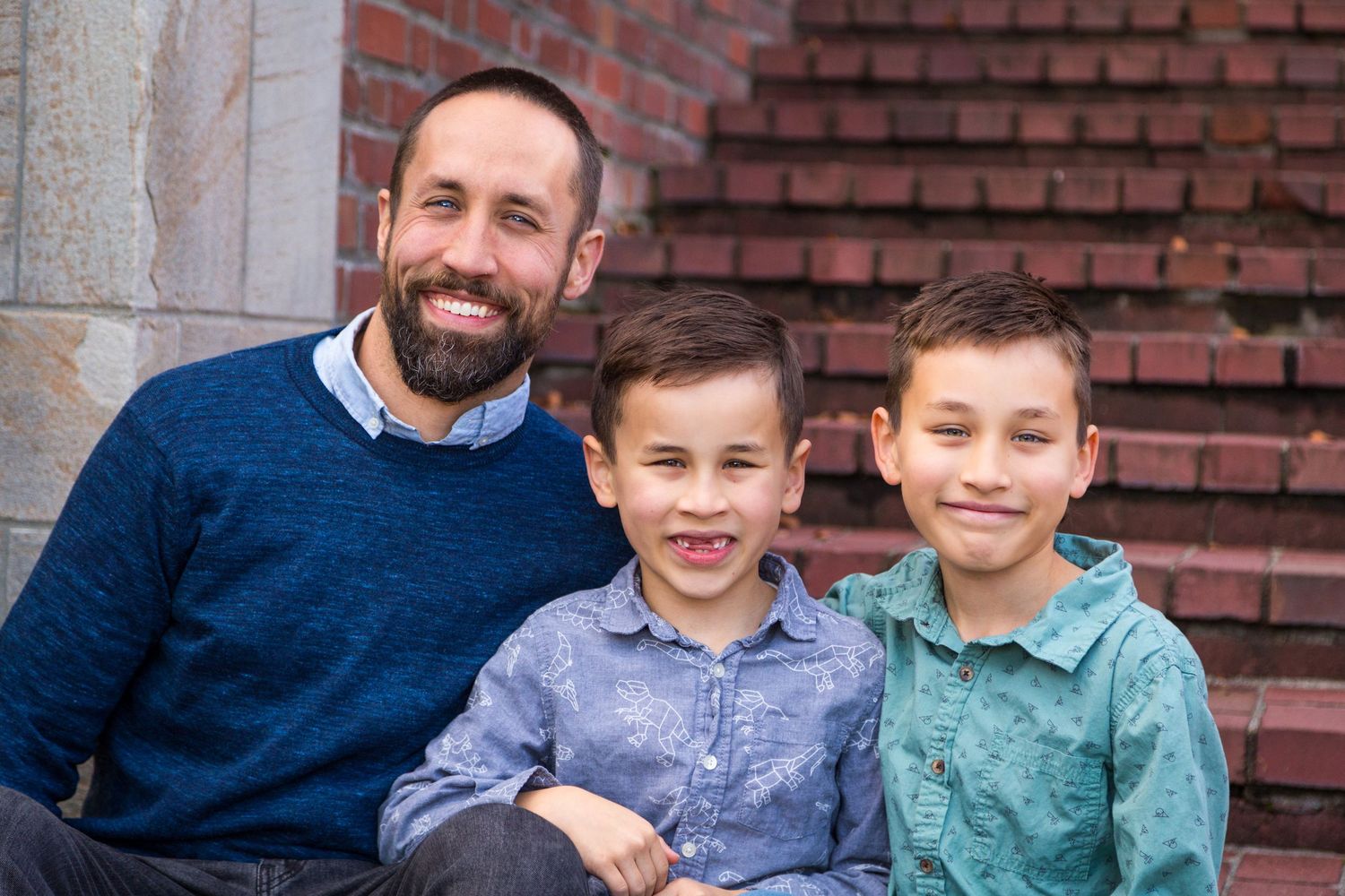 A cheerful parent sits with two young boys on brick stairs while wearing a blue sweater.
