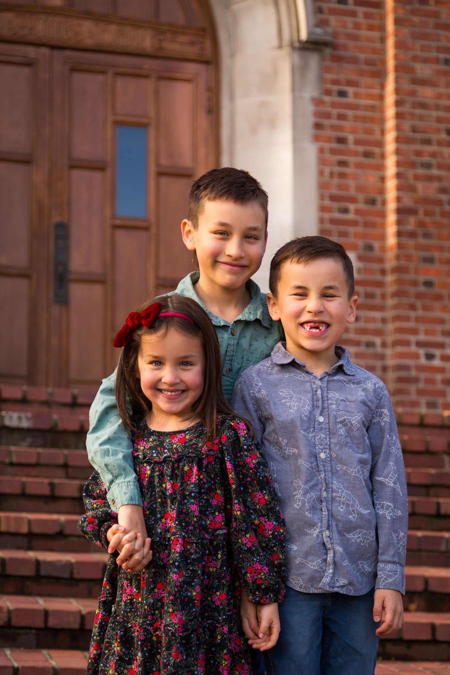Three children stand together smiling on brick steps outside a wooden door.