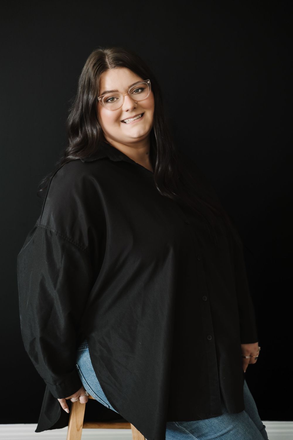 Person wearing glasses and black sweater poses against dark background while sitting on stool.
