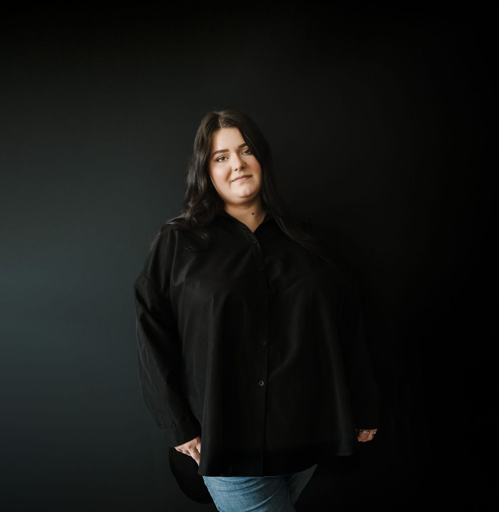 Studio portrait of someone in a black button-up shirt standing against a dark green background.