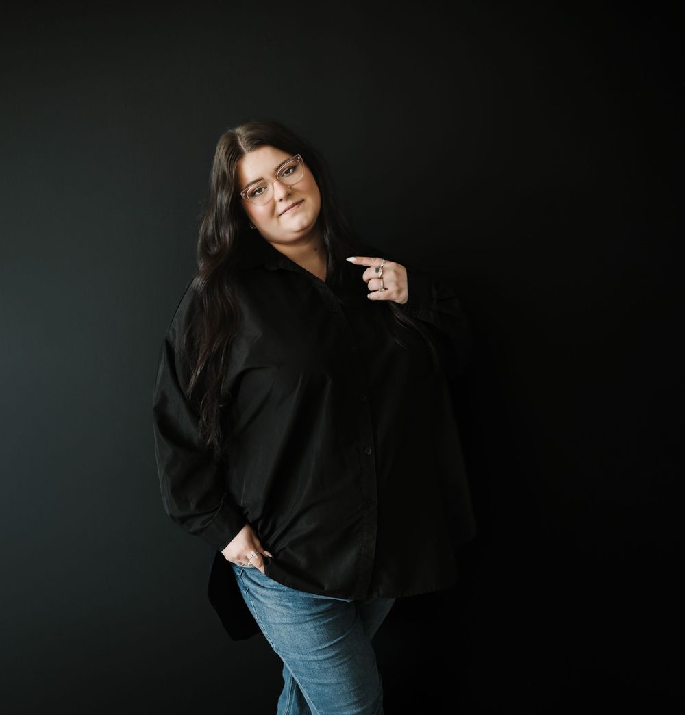 A person wearing a black oversized sweater and blue jeans poses against a dark backdrop in a casual studio portrait.