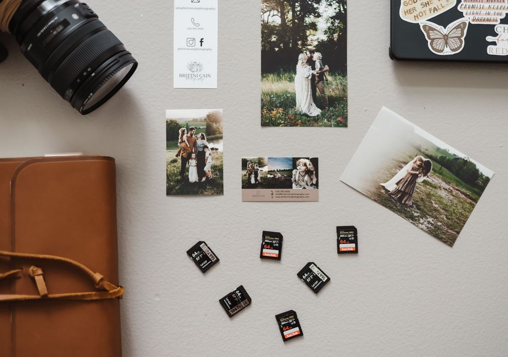 Flatlay showing camera lens, leather journal, memory cards and wedding photography prints arranged on white surface.