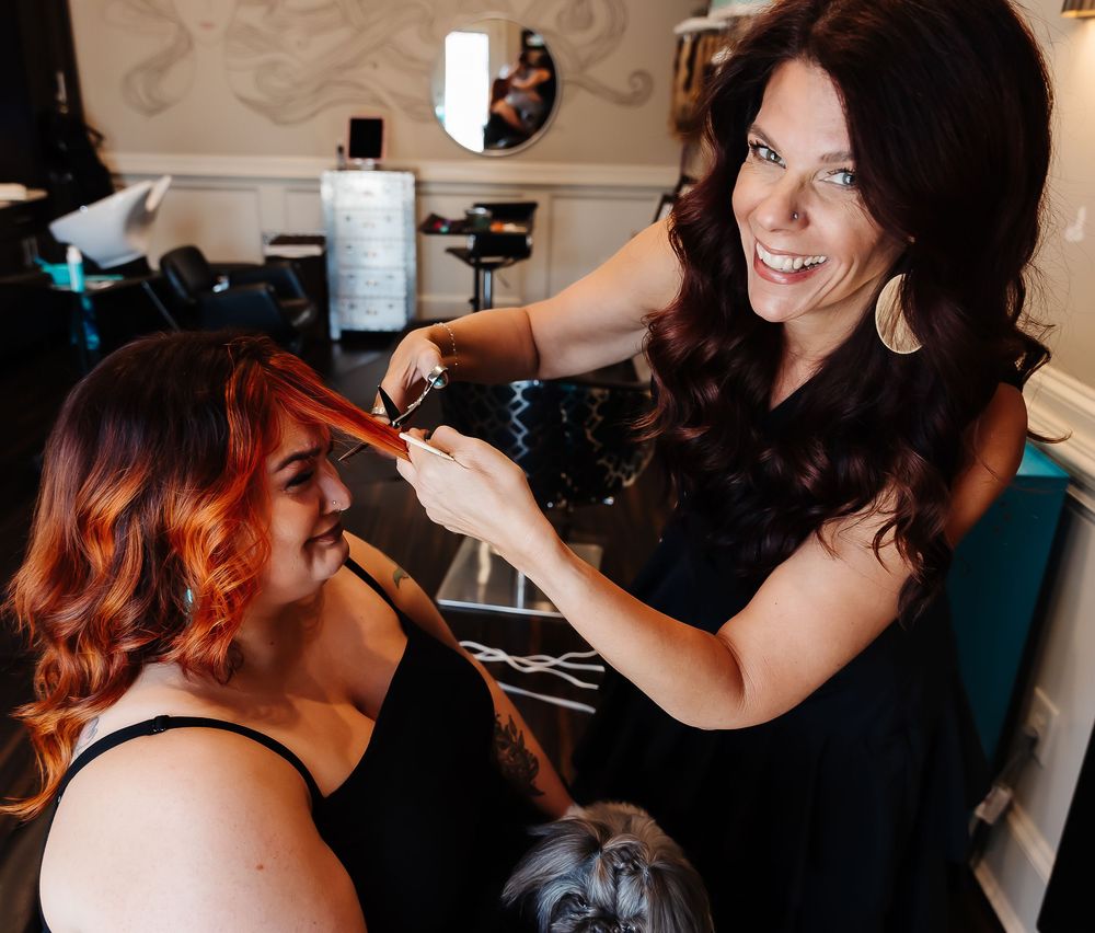 Hair stylist with dark hair works on client's vibrant red-tinted curls at a salon station.