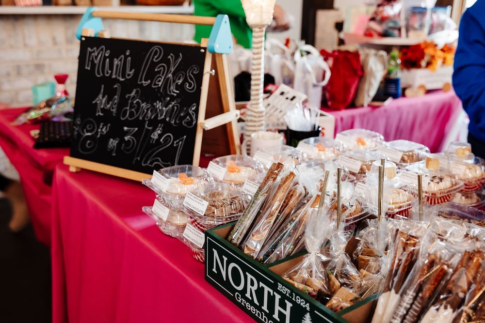 A vendor booth at a craft fair displays homemade treats and baked goods on a red tablecloth with a chalkboard sign.