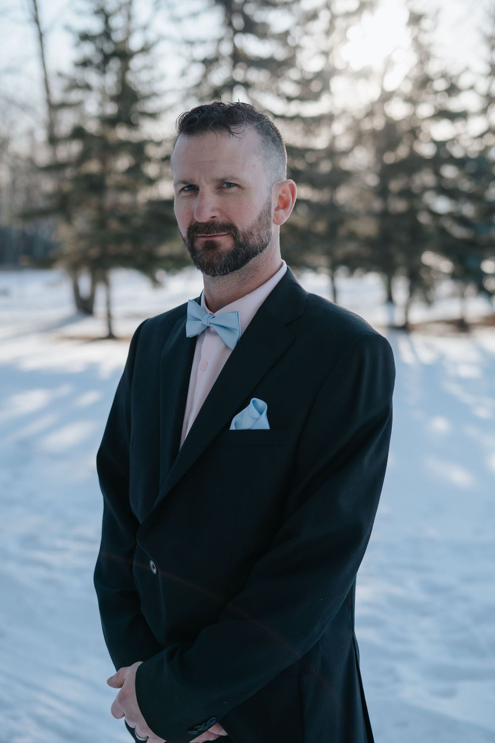 A person in a black tuxedo with light blue pocket square stands in a snowy landscape.