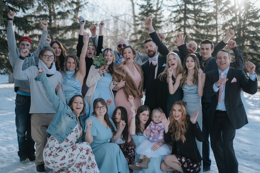 Joyful wedding party celebrating in snow with evergreen trees in background.