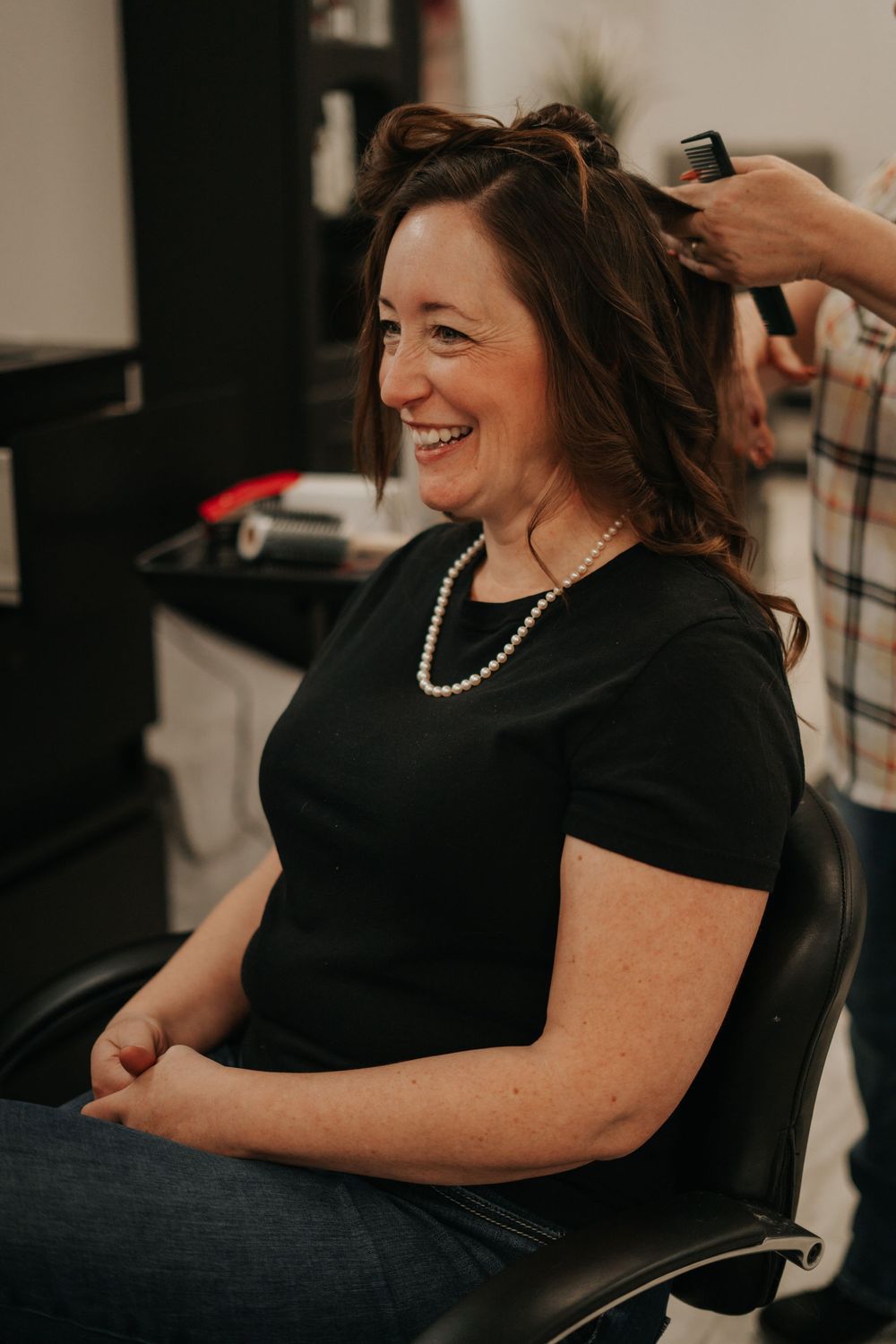 A person wearing pearls and black top gets their hair styled.