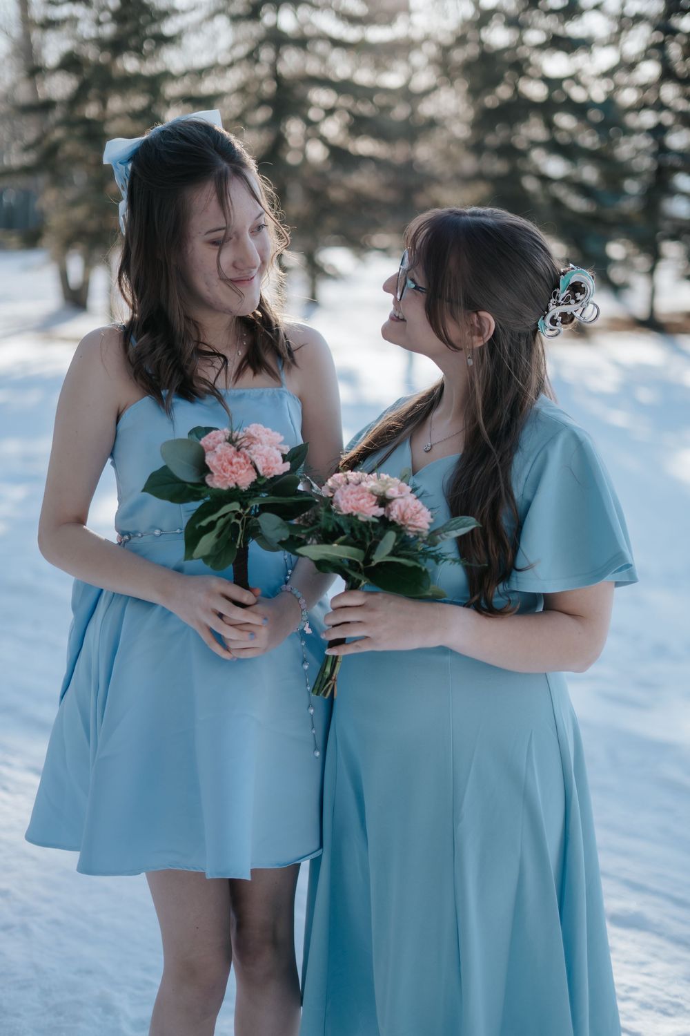 Two bridesmaids in light blue dresses hold pink bouquets in a snowy setting.