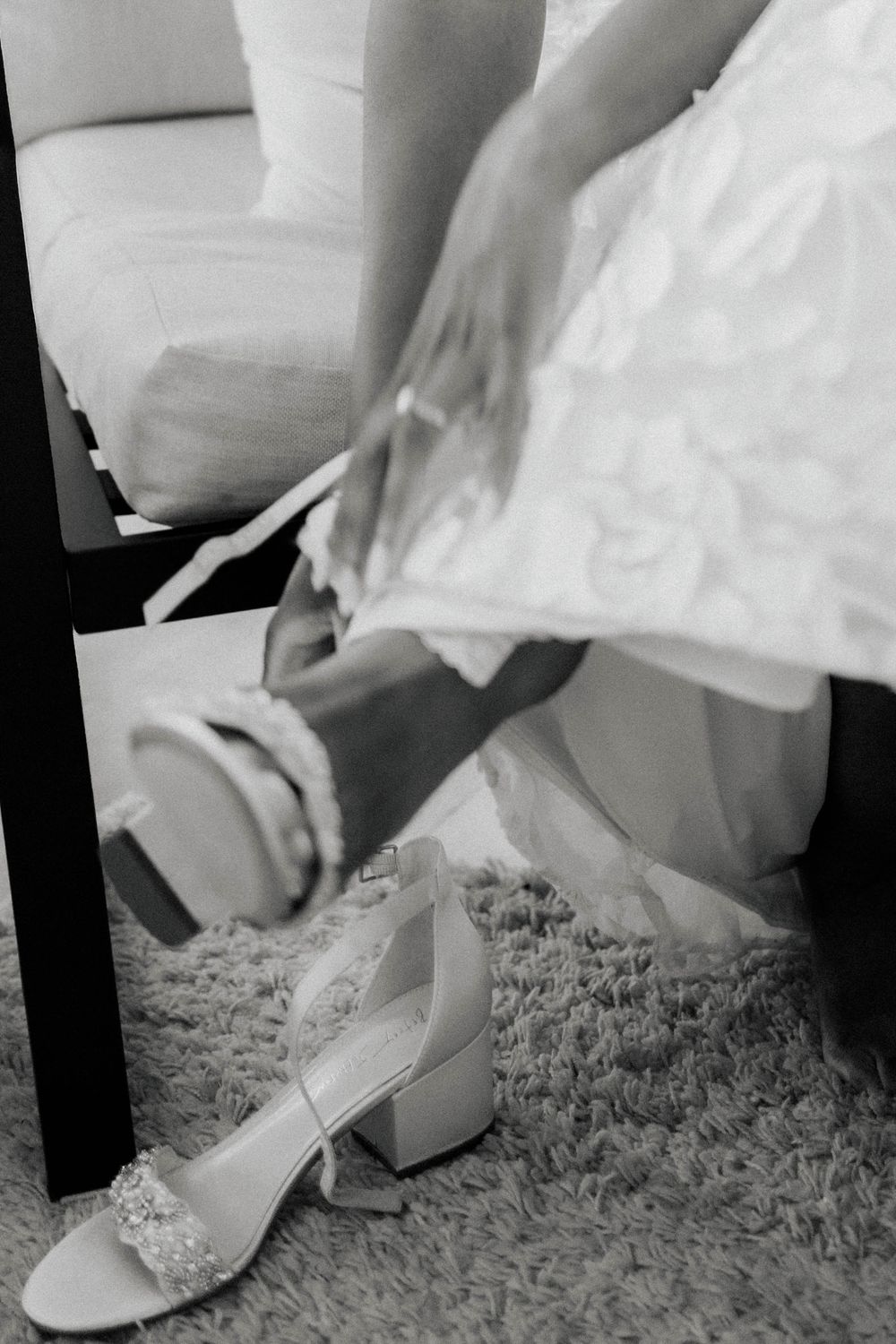 Black and white close-up of bride putting on jeweled block heel wedding shoes.