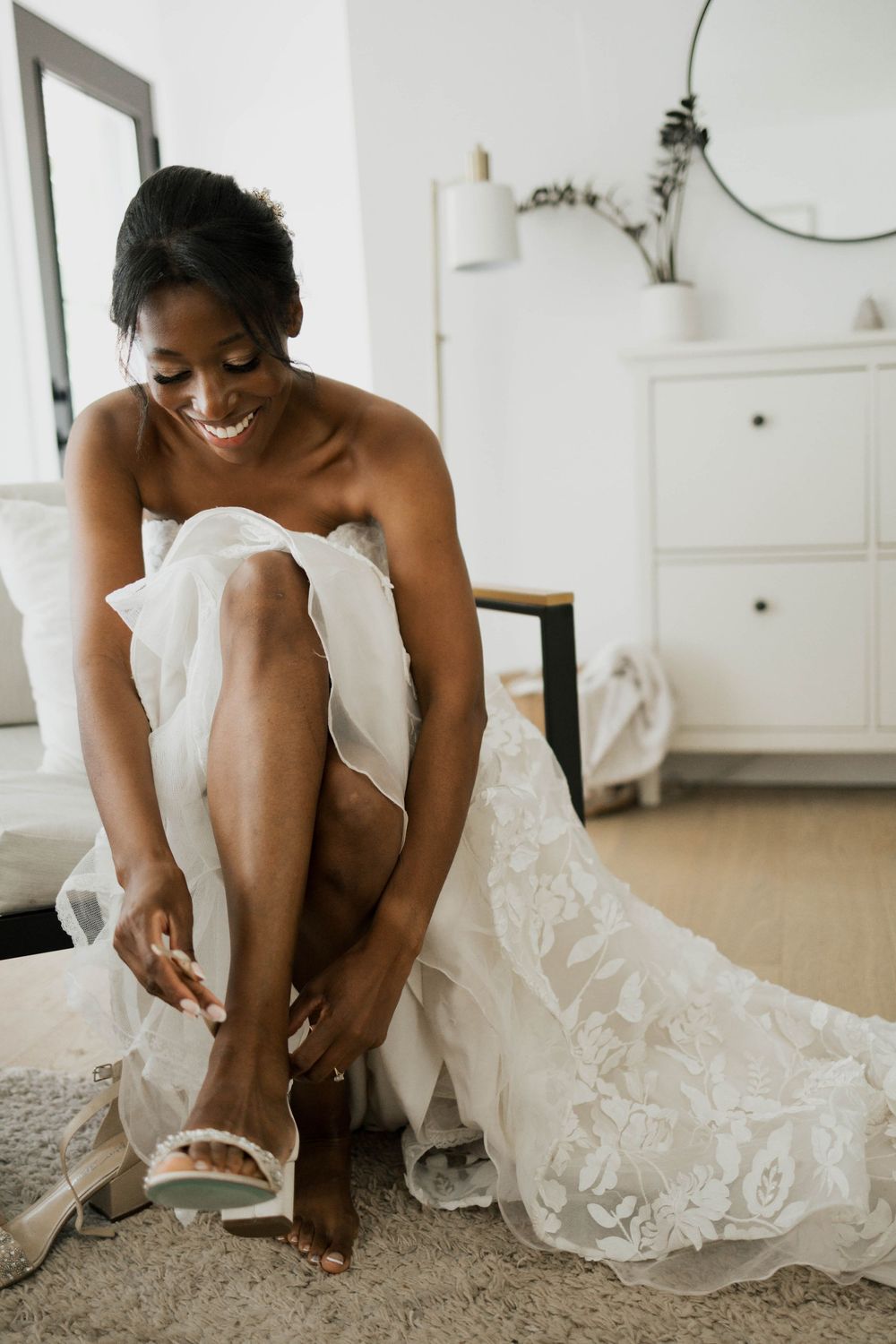 Radiant Black bride in a lace wedding gown smiles while putting on her bridal heels before the ceremony.