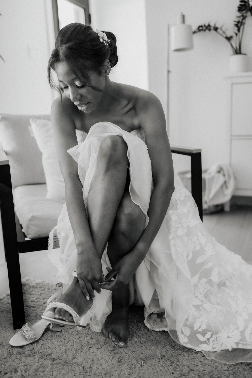 Black bride in a strapless lace wedding gown fastens her bridal sandals while seated before her wedding.