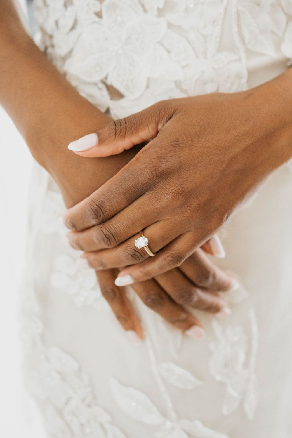Close-up of bride's hands showing diamond solitaire engagement ring on lace wedding gown.