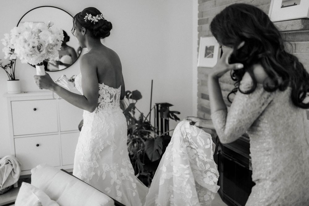 Black bride in a lace wedding gown holds her bouquet while a bridesmaid sits nearby during bridal prep.