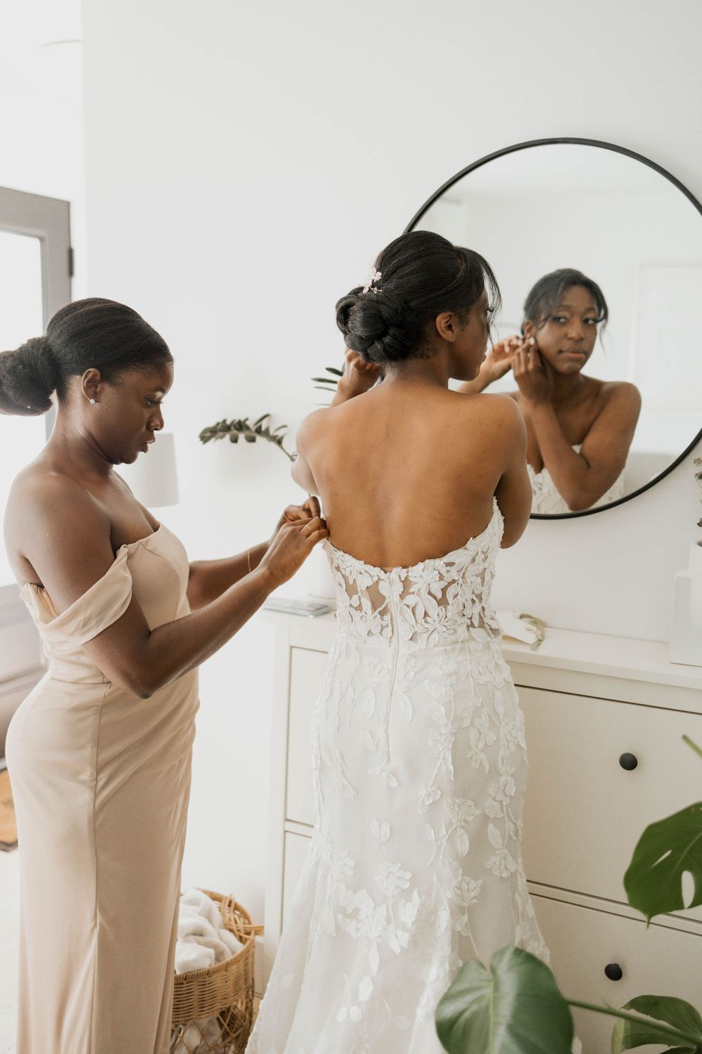 Bridesmaid in a champagne gown buttons the lace back of the Black bride's wedding dress before the ceremony.