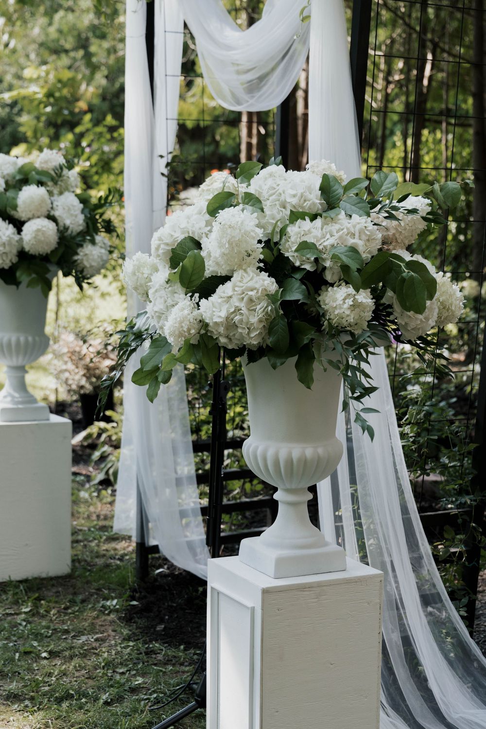 White hydrangea floral arrangements in urn vases at outdoor wedding ceremony altar.