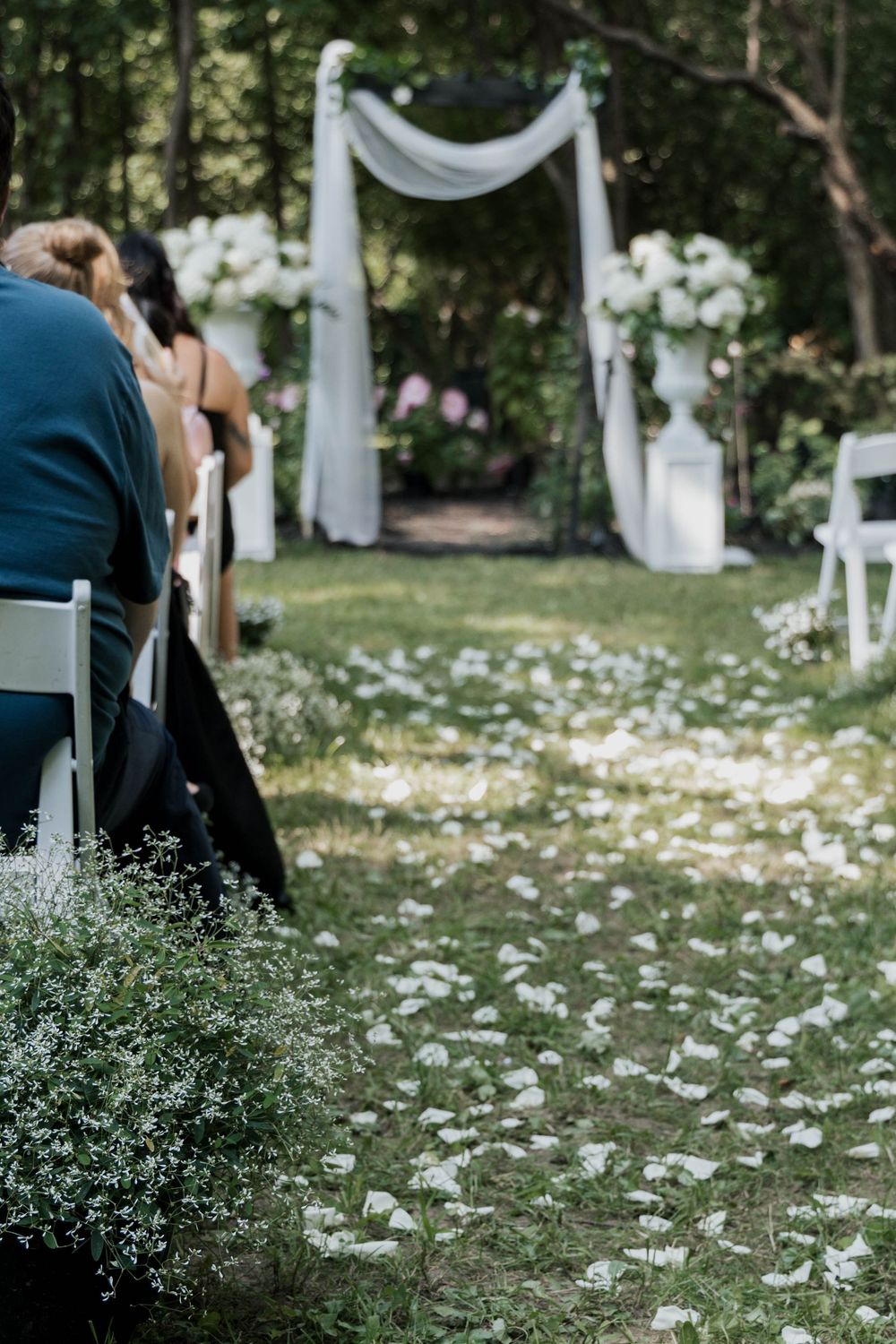 Elegant outdoor wedding aisle with white flower petals leading to a draped floral arch in a lush forest.