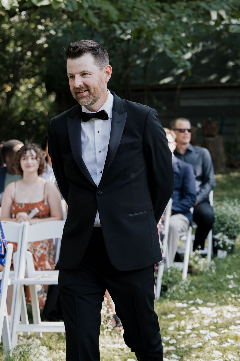 Handsome groom in black tuxedo and bow tie smiling at outdoor wedding ceremony with seated guests behind.