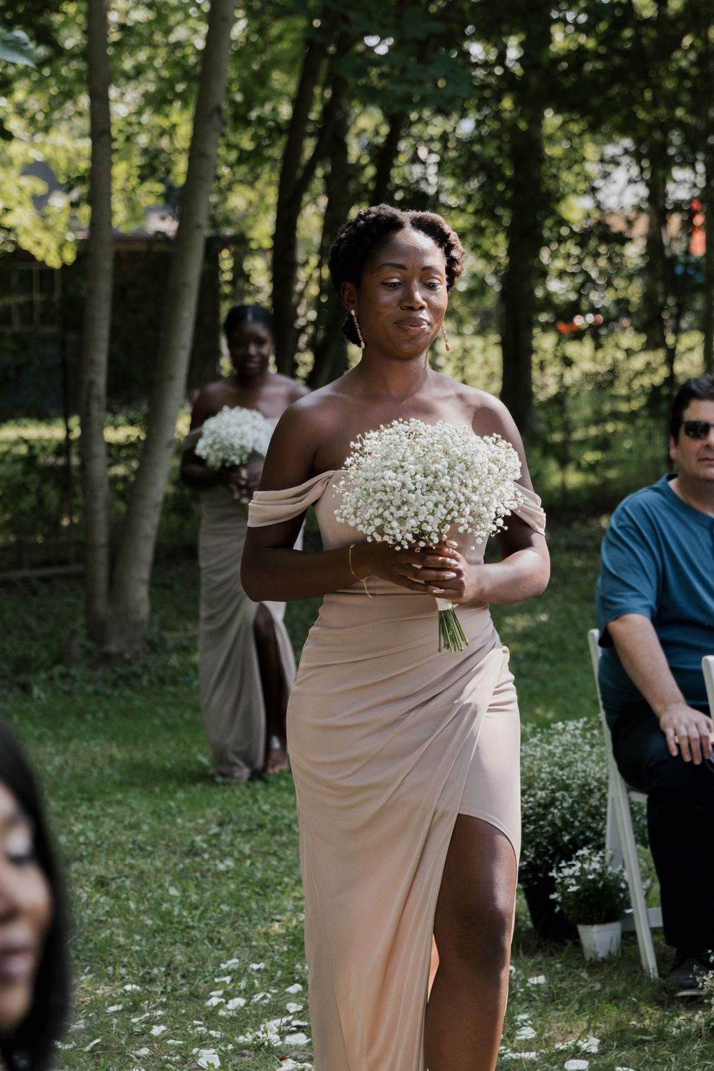 Bridesmaid in champagne off-shoulder gown carrying baby's breath bouquet down aisle.