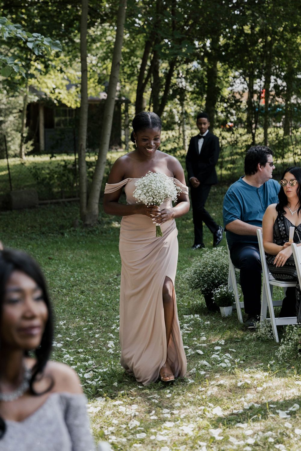 Smiling bridesmaid in champagne off-shoulder gown carrying baby's breath bouquet walking down outdoor wedding aisle.