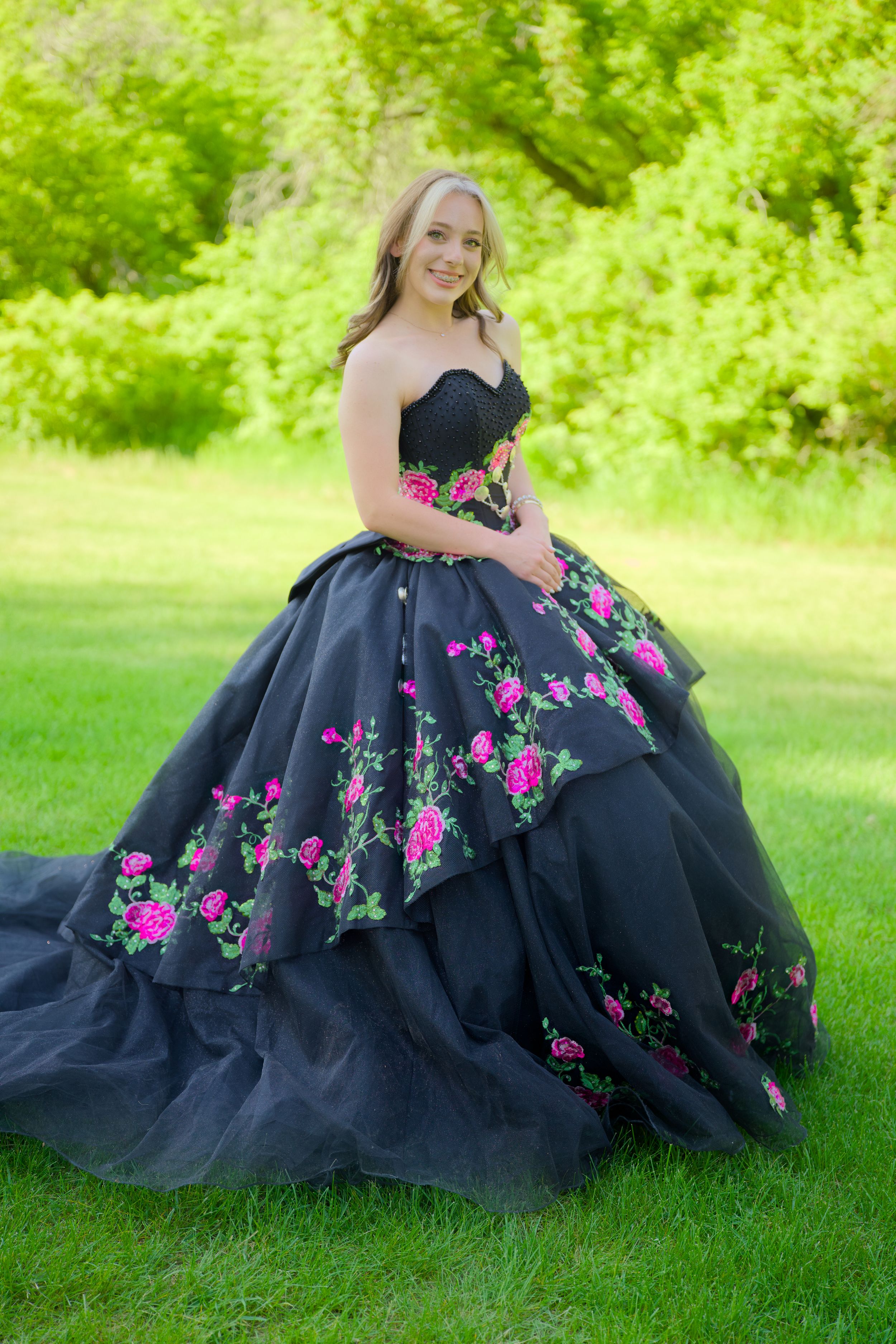 Elegant black ballgown with pink rose embellishments photographed in a lush green outdoor setting.